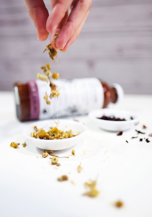 Woman's hand sprinkling camomile flowers into a white dish with a bottle of Thrive Remedies Calm flavour in the background